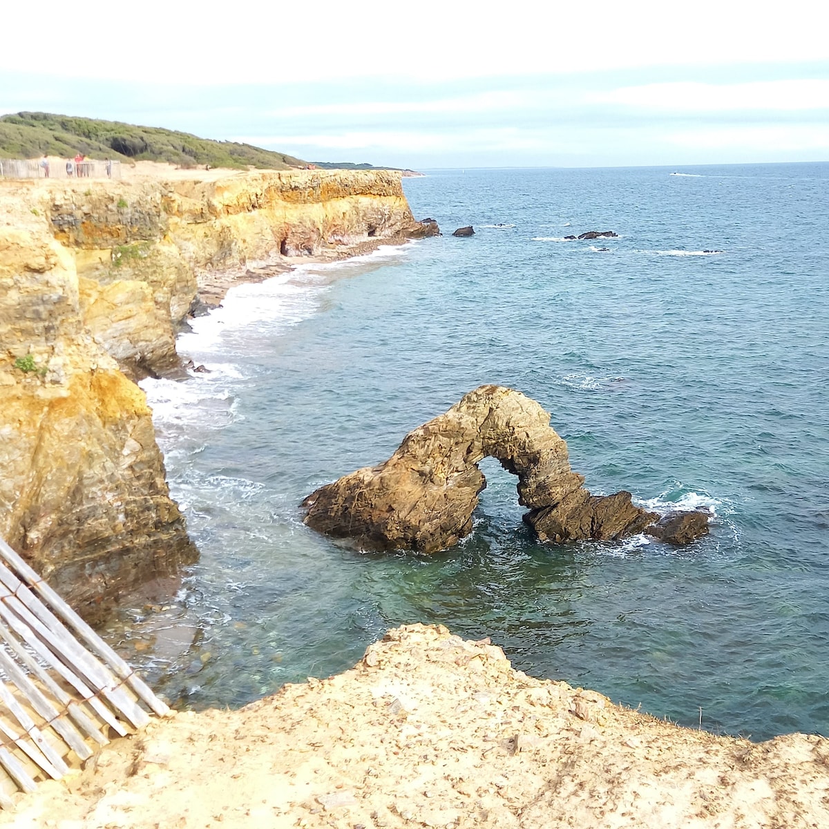 A coastal landscape features a natural rock formation arching over the sea, surrounded by rugged cliffs. Gentle waves lap against the shore, while a portion of the coastline extends into the distance, offering a serene view of the horizon.