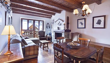 The living area features wood-beamed ceilings and a blend of comfortable seating. A rustic dining table accommodates six, while a cozy sofa and armchairs invite relaxation. Natural light filters through the windows, highlighting the decorative art on the walls and a fireplace positioned thoughtfully.