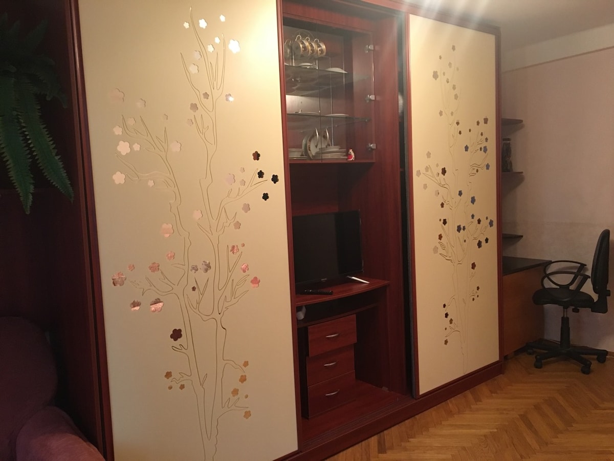A modern entertainment center features decorative doors adorned with floral designs. Inside, a small television is visible, complemented by shelves displaying decorative items. A workspace with a chair is situated to the right, while a soft seating area is present to the left.