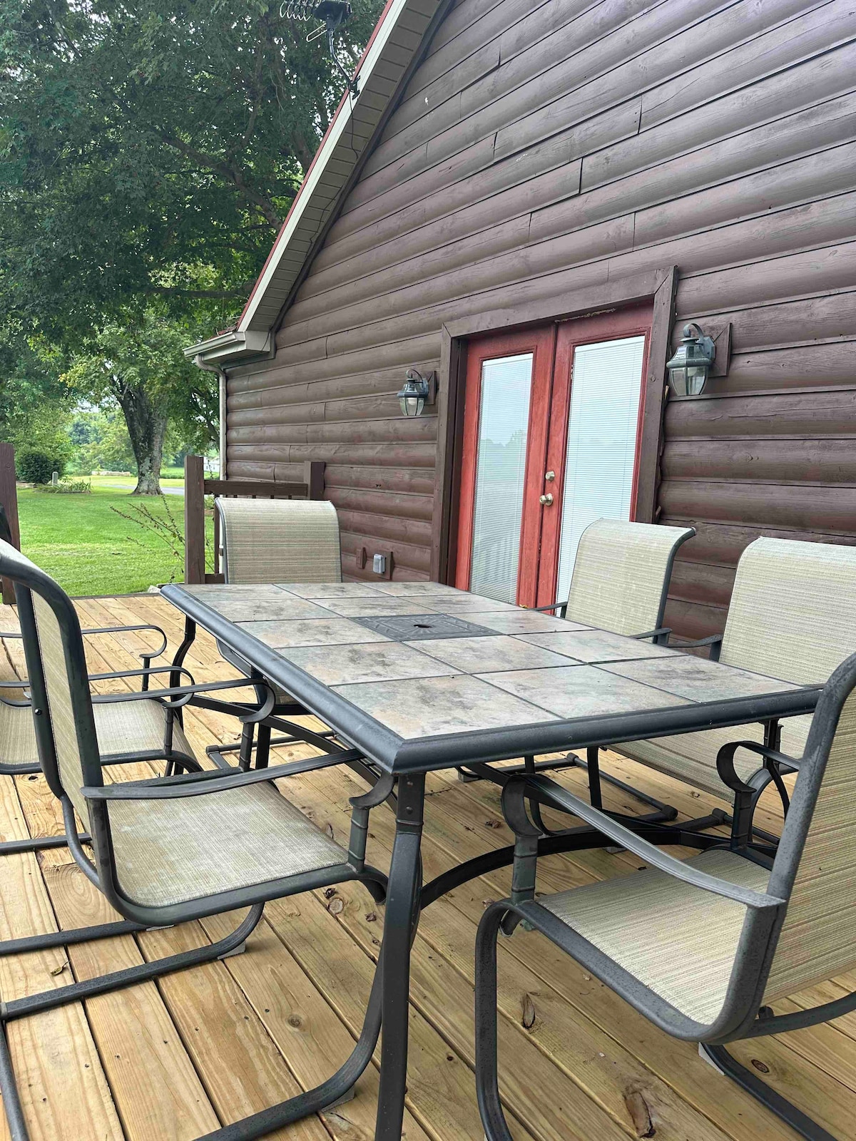 An outdoor dining area features a rectangular table surrounded by six light-colored chairs on a wooden deck. The cabin's brown wooden exterior is visible in the background, complemented by double doors that provide access to the interior. Lush greenery is seen beyond the deck.