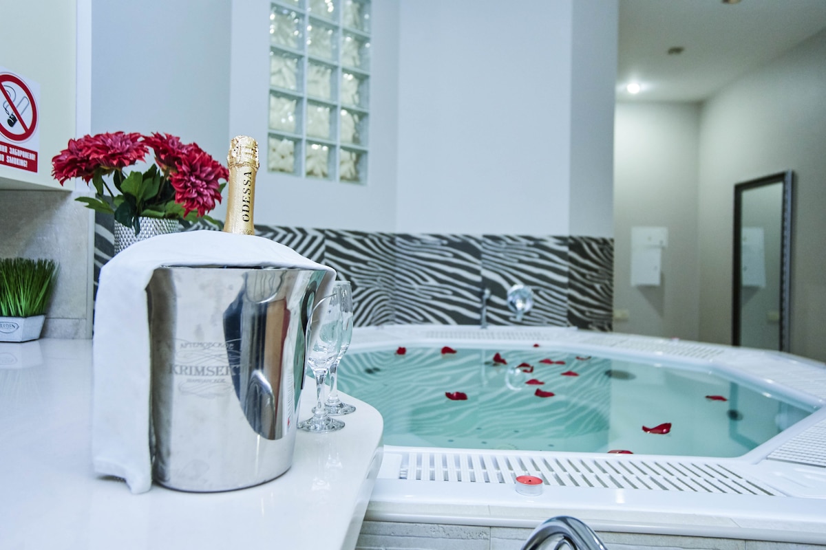 A well-appointed area features a large spa pool adorned with rose petals. A silver champagne bucket and two elegant glasses are positioned on the nearby marble counter. The walls are decorated with a modern zebra-patterned design, complementing the contemporary ambiance.