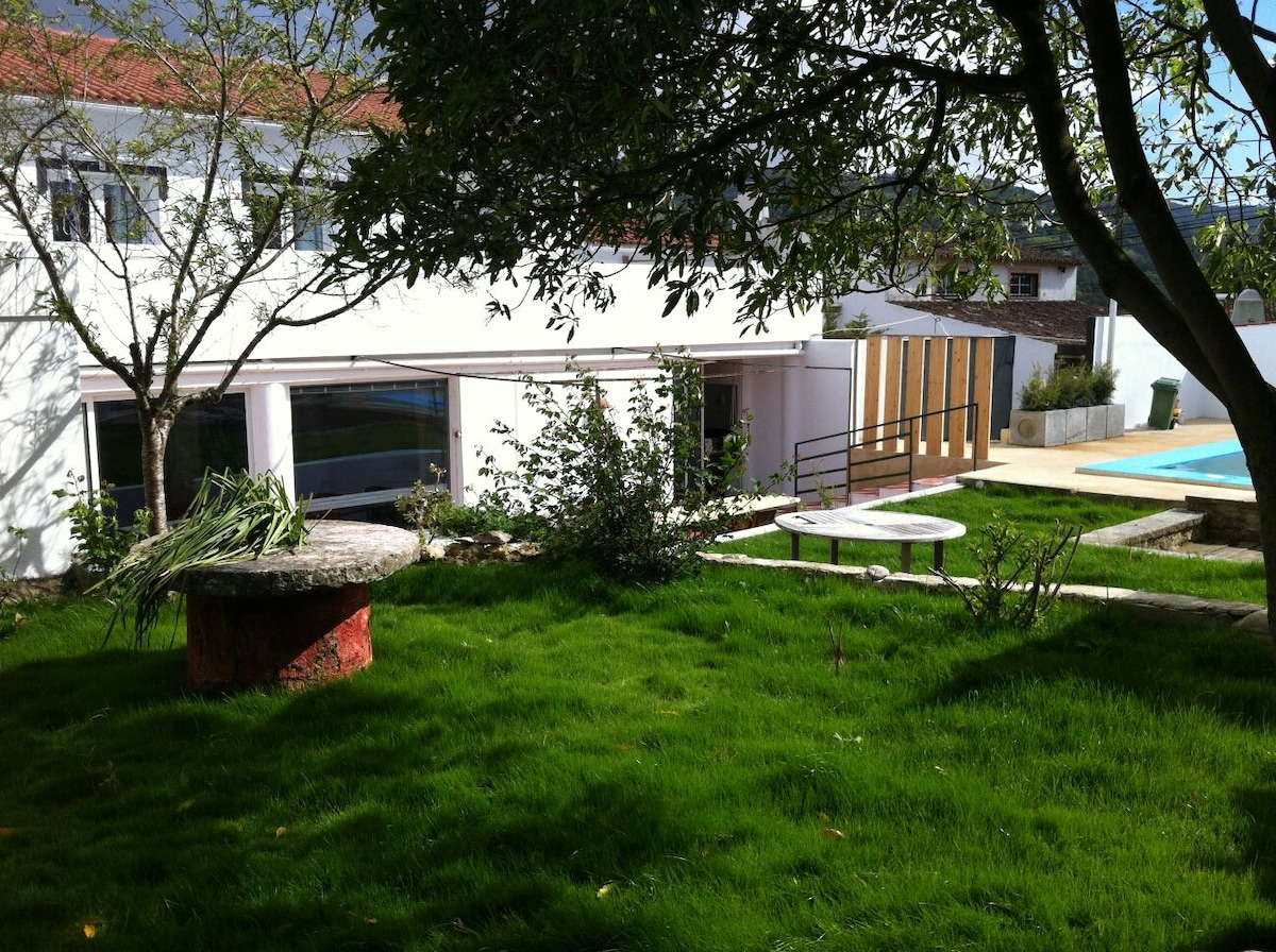 A landscaped garden featuring lush green grass is visible, with a round stone table and a rustic stone structure. Sunlight filters through the trees, highlighting the two private terraces and the shared swimming pool in the background, which is framed by a contemporary building.