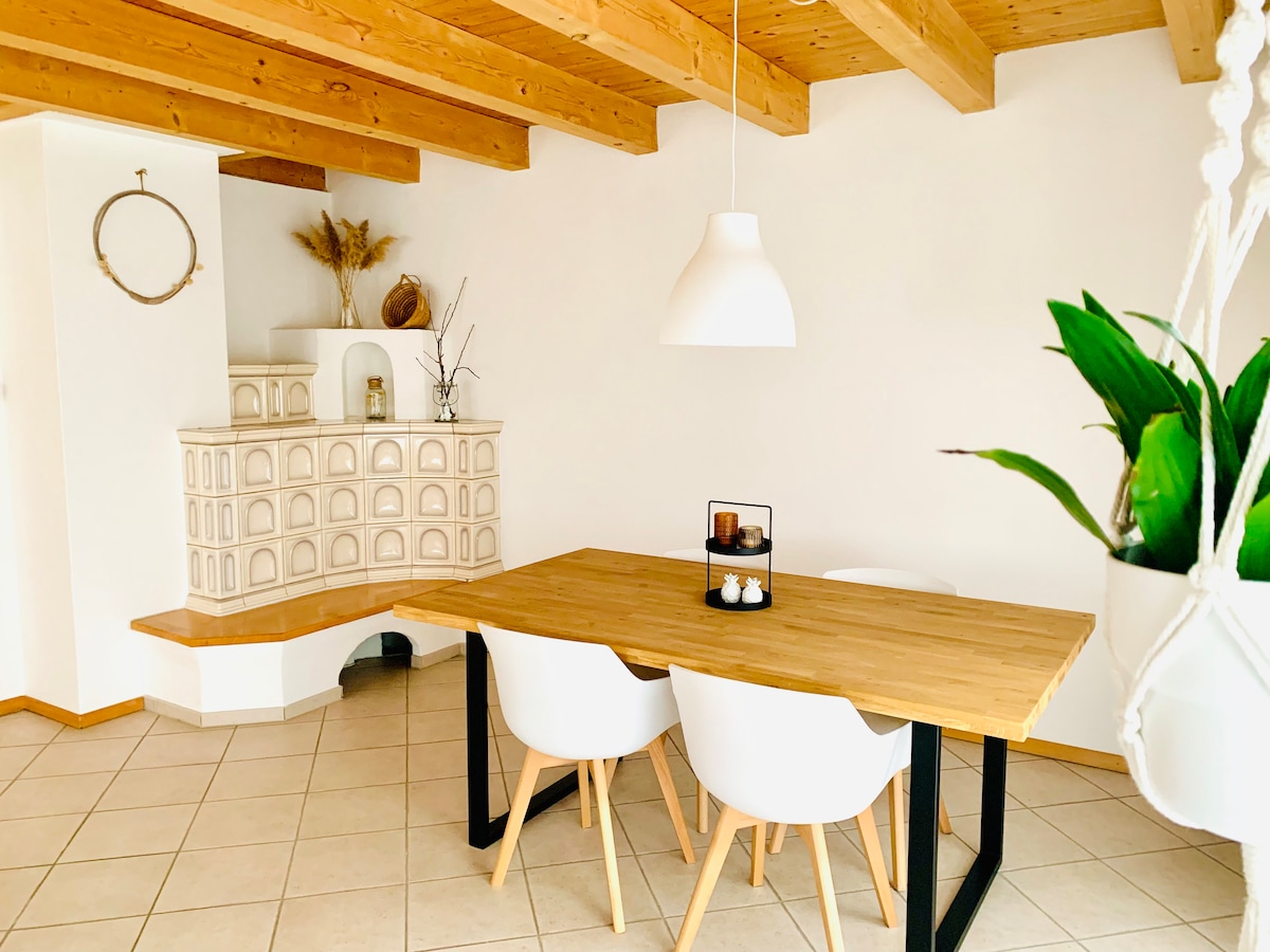 A dining area is shown with a wooden table paired with four white chairs. A white pendant light hangs overhead while a tiled stove is positioned against the wall. Decorative plants and natural accents add a touch of warmth to the space.
