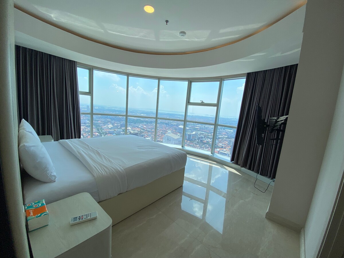 A contemporary bedroom offers expansive windows that showcase panoramic city views. The floor is polished, reflecting natural light that fills the space. A spacious bed is positioned against a curved wall, complemented by dark curtains and a bedside table.