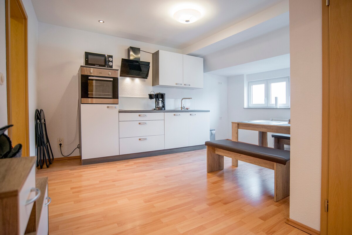 A modern kitchen is presented with sleek cabinetry and essential appliances, including a microwave and coffee maker. A spacious wooden dining table is positioned near large windows, providing ample natural light. A bench is situated alongside the table, enhancing the functionality of the space.