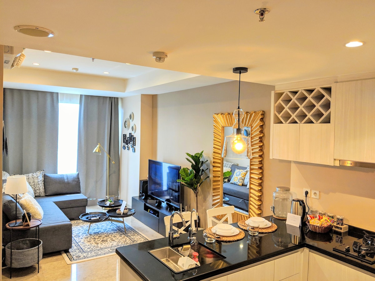 An open-plan living space features a modern kitchen with dark countertops and a dining area set for four. A comfortable sofa is positioned facing a flat-screen TV, with decor elements such as plants and a mirror enhancing the ambiance. Natural light fills the room.