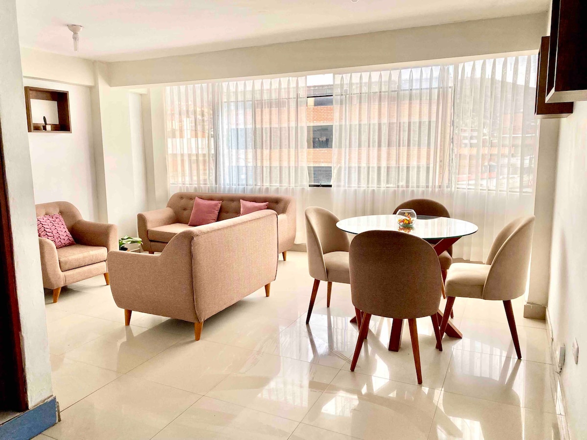 The bright living area features large windows with sheer curtains, allowing natural light to fill the space. A round dining table with upholstered chairs sits next to a comfortable sofa set, creating a welcoming environment for relaxation and meals.