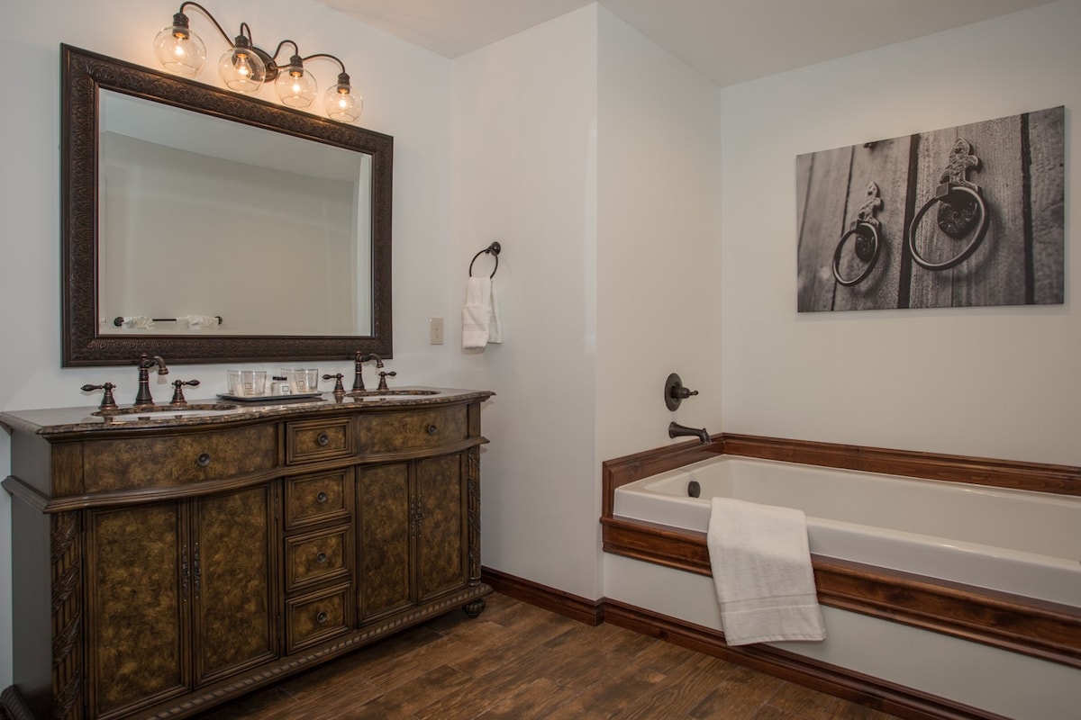 A stylish bathroom features a dark wood vanity with a large framed mirror above. A modern bathtub is positioned beside it, with a towel hanging on the side. A monochromatic photograph of bicycle wheels is displayed on the wall, complementing the neutral color scheme.