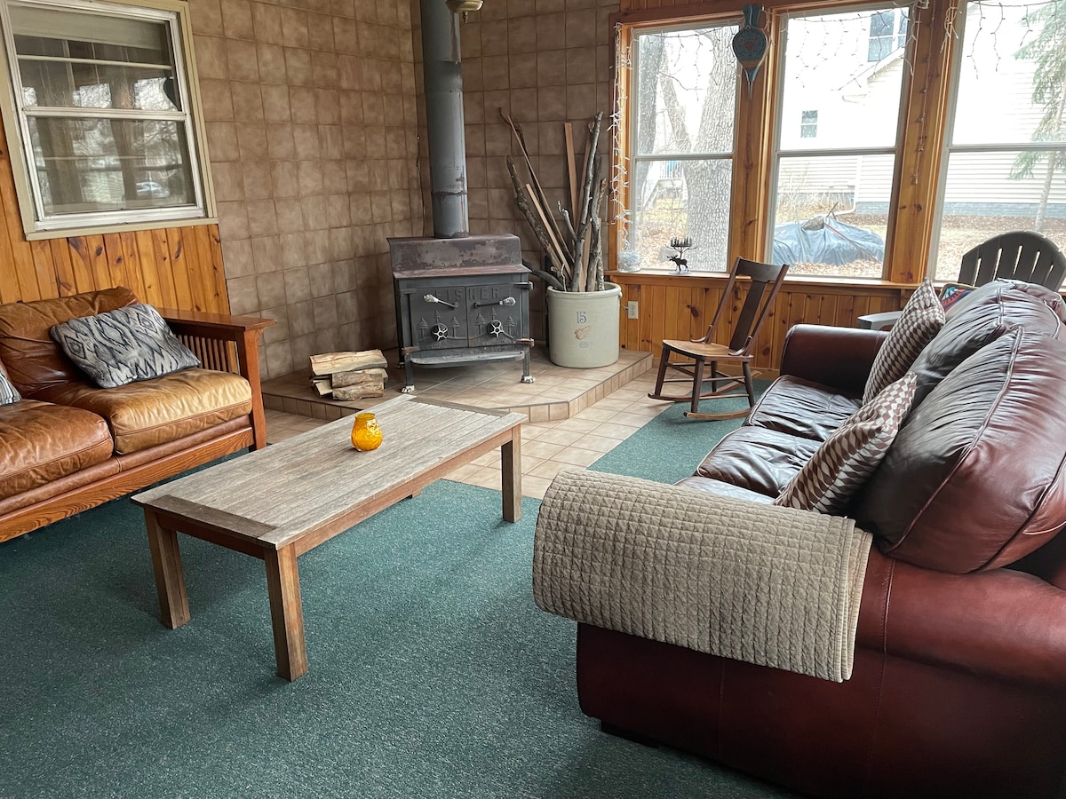 A cozy porch is presented with a rustic wooden coffee table at the center, flanked by a leather sofa and a wooden loveseat. A wood-burning stove is positioned against the wall, surrounded by decorative logs. Natural light is filtered through large windows, enhancing the inviting space.