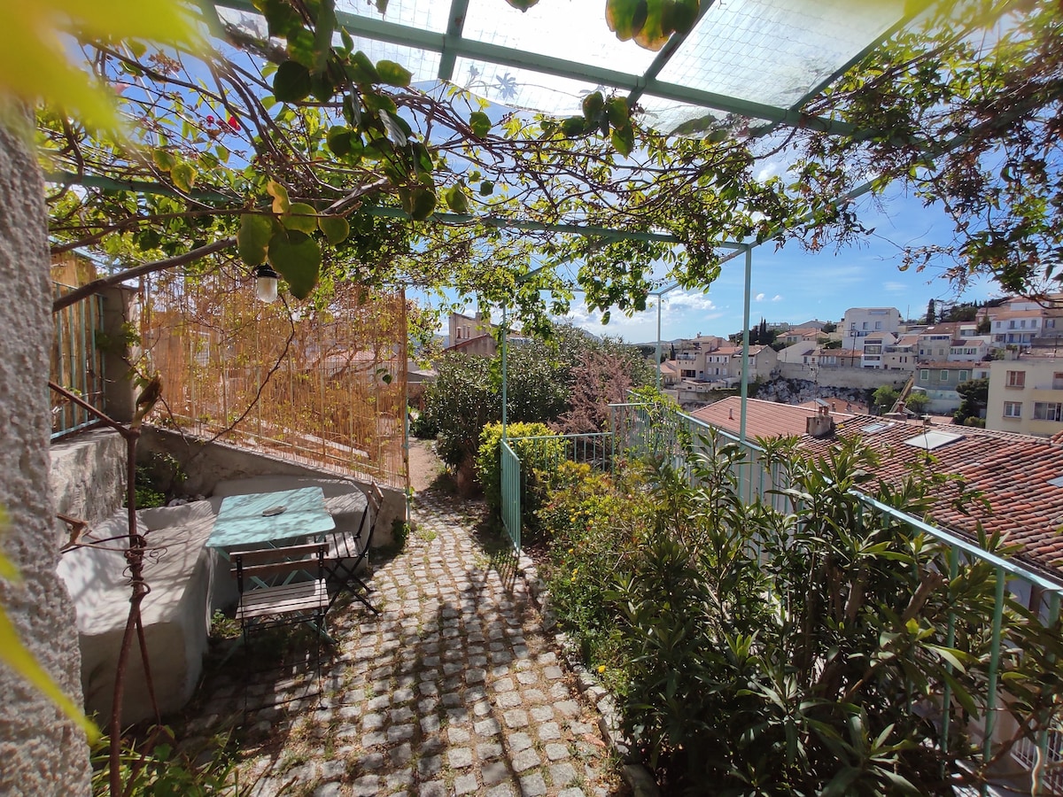 A garden terrace is surrounded by lush greenery, featuring a cobblestone path. A small table and chairs are positioned under a trellised canopy, offering shade. Houses can be seen in the background, while sunlight illuminates the vibrant space.
