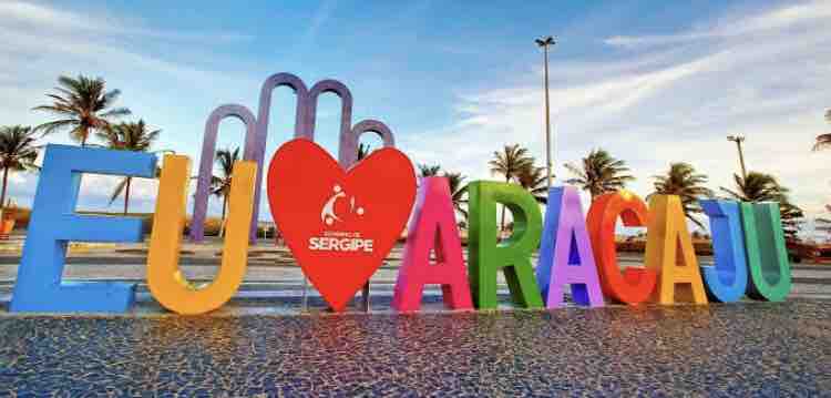 A colorful sign displaying the phrase 'EU AMO ARACAJU' is set against a backdrop of palm trees and a clear sky. The vibrant letters, in various colors, create a welcoming atmosphere. The ground is paved with stone, reflecting the surrounding natural beauty.