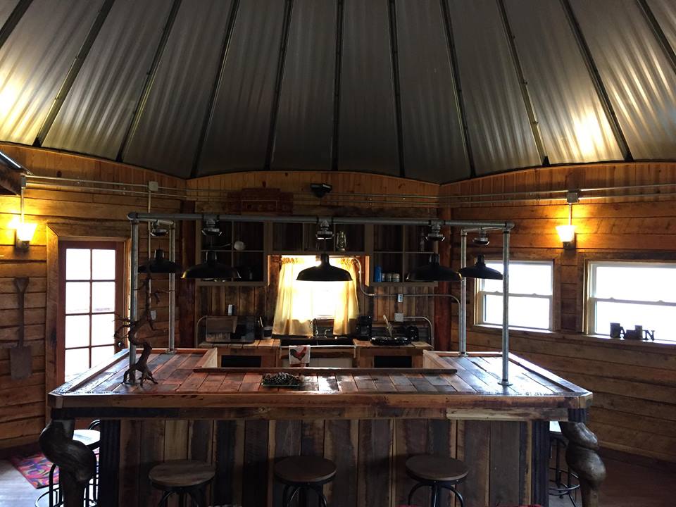 An open kitchen area features a large U-shaped bar surrounded by several stools. The space is illuminated by pendant lights and showcases rustic wooden walls. Natural light streams in through multiple windows, highlighting the unique design and warm atmosphere of the yurt.