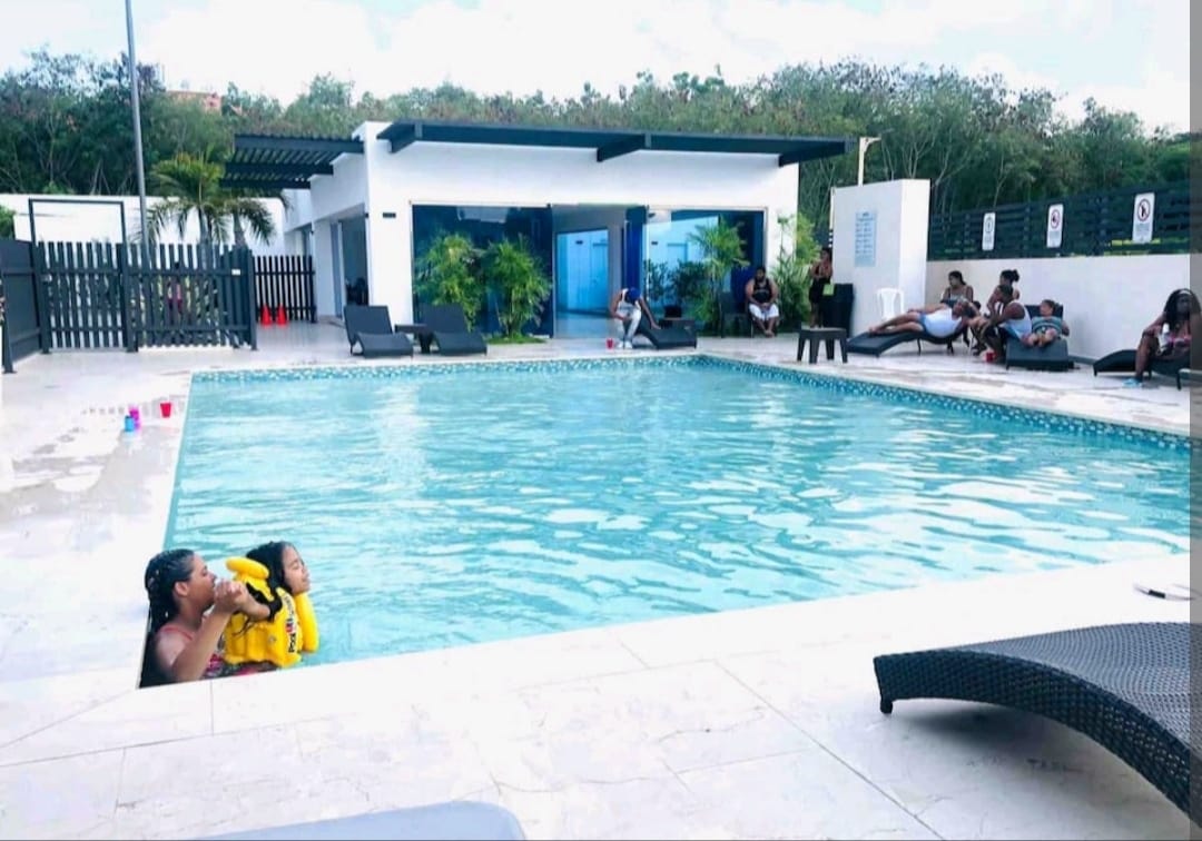 A spacious swimming pool is surrounded by lounge chairs. Several guests are seen enjoying the water and relaxing poolside. The area is bordered by lush greenery and features a modern building in the background, providing a serene atmosphere for leisure.