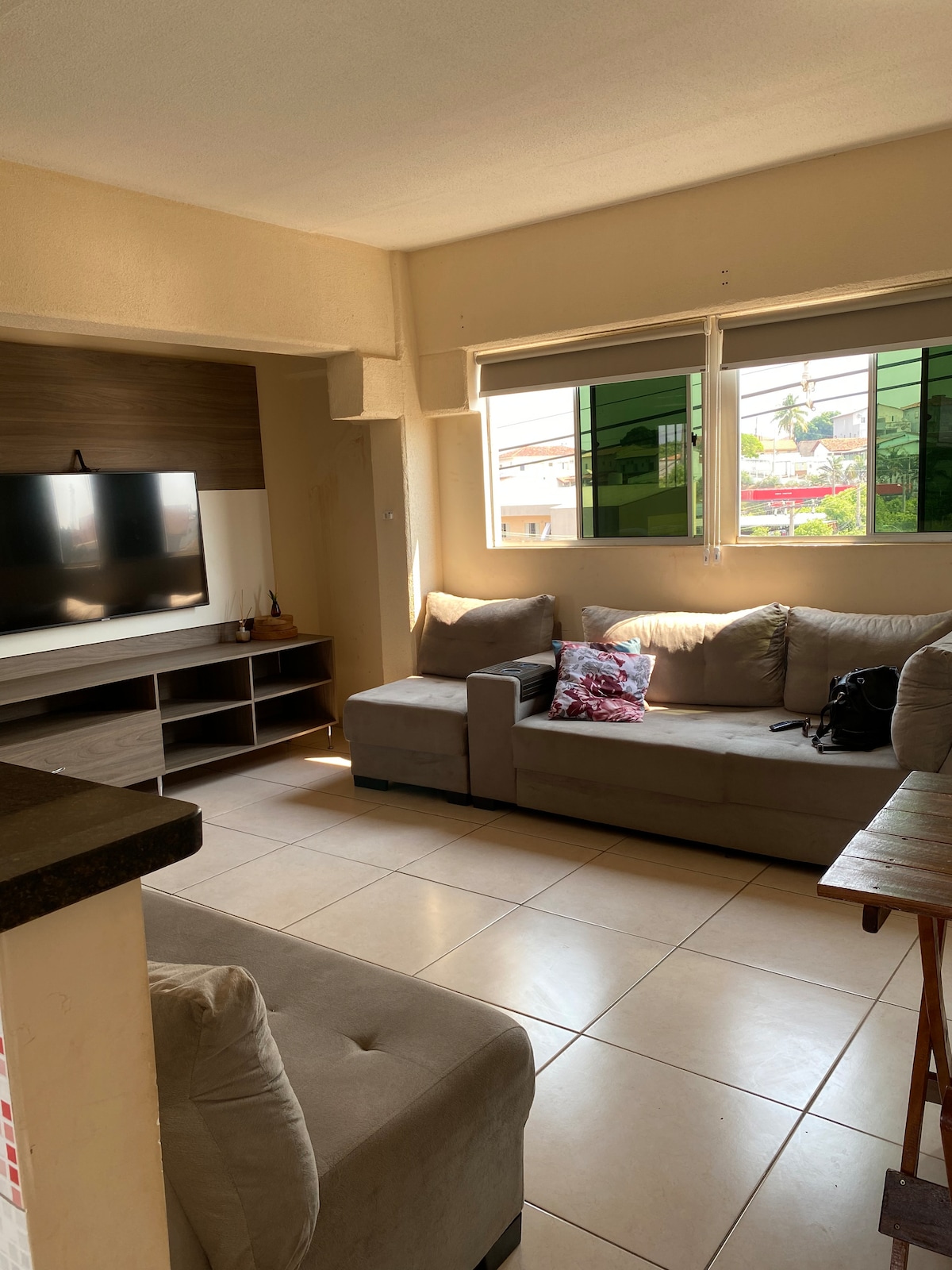 A living area is presented with soft beige seating arranged around a flat-screen television. Two large windows allow natural light to fill the space, revealing glimpses of greenery outside. A small table can be seen in the foreground, enhancing the cozy layout.