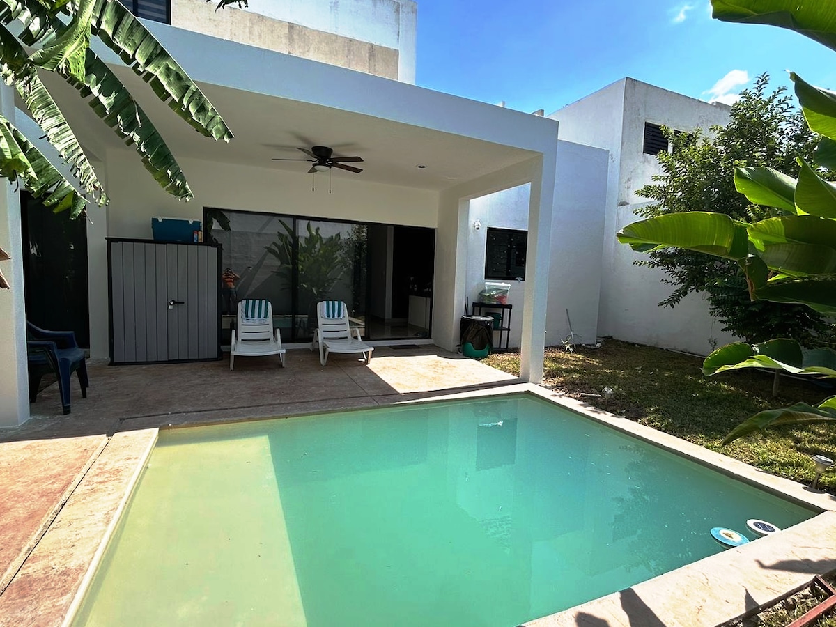 A private outdoor area features a tranquil pool surrounded by a spacious patio. Two lounge chairs are positioned nearby under a shaded area. Lush greenery and banana plants add a touch of nature to the setting, creating a relaxing environment.