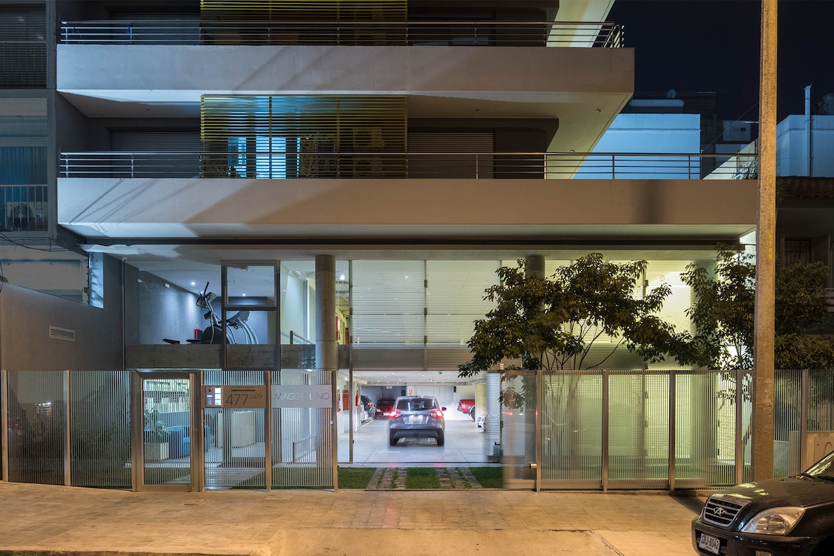 The modern building facade is illuminated at night, showcasing its sleek design. The entrance features large glass panels, allowing visibility into the lobby area. Vehicles are seen entering the parking area, surrounded by greenery that lines the pathway.