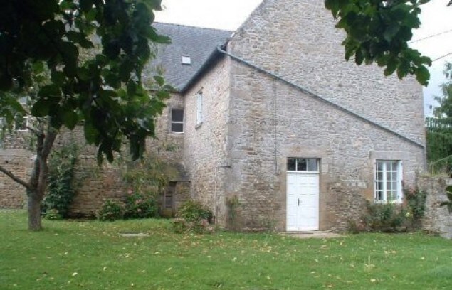 The exterior of a stone house features a simple white door and multiple windows, surrounded by a well-maintained green lawn. Trees frame the image, enhancing the natural surroundings.