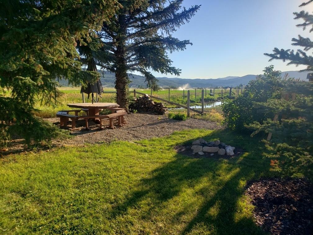 A shaded outdoor area features a wooden picnic table surrounded by natural landscaping. Nearby, neatly stacked firewood is positioned against a backdrop of lush greenery and a gentle stream. The distant mountains provide a scenic horizon under a clear blue sky.