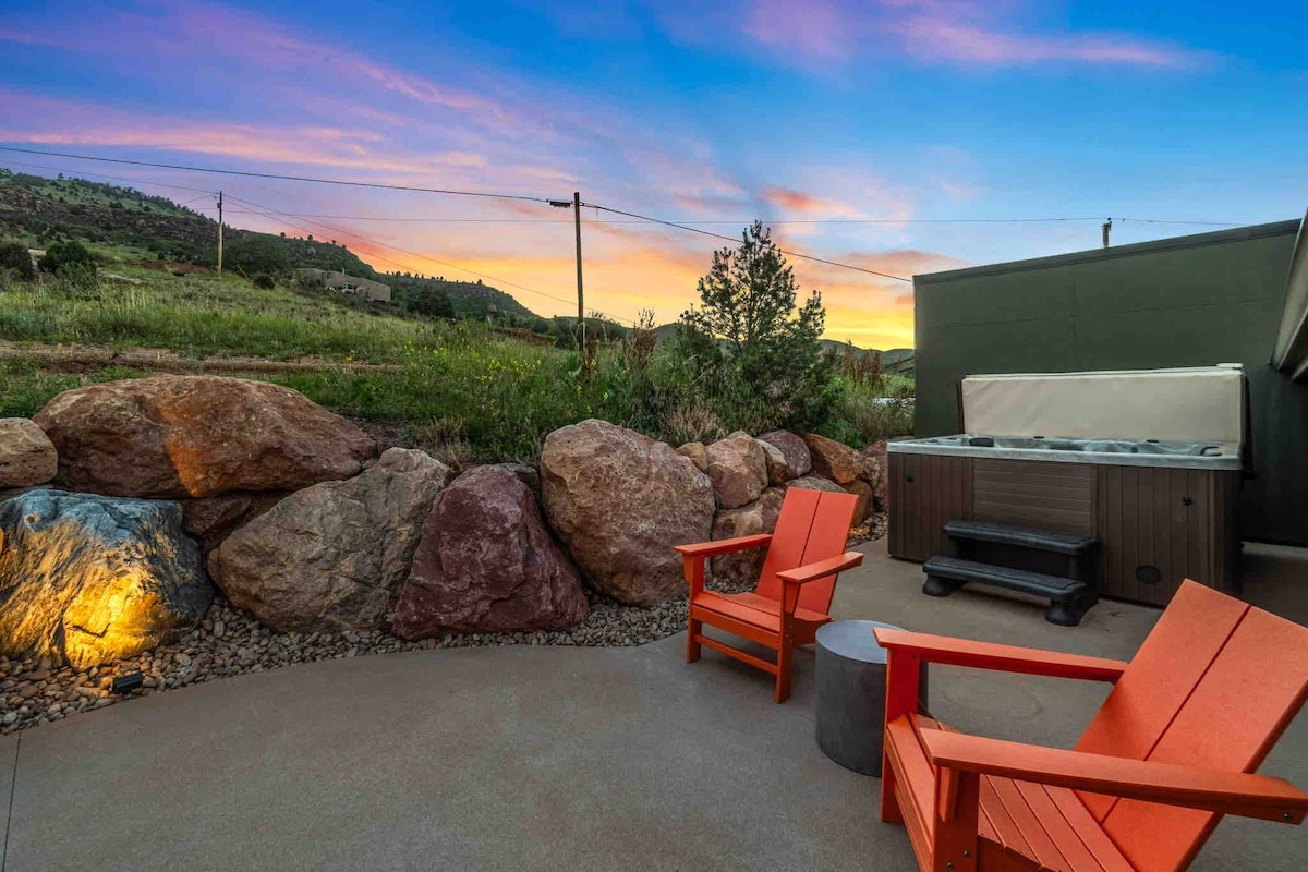 An outdoor patio area features two orange adirondack chairs positioned beside a hot tub, with natural rock formations lining the space. A scenic view of the surrounding landscape is enhanced by a colorful sunset sky.