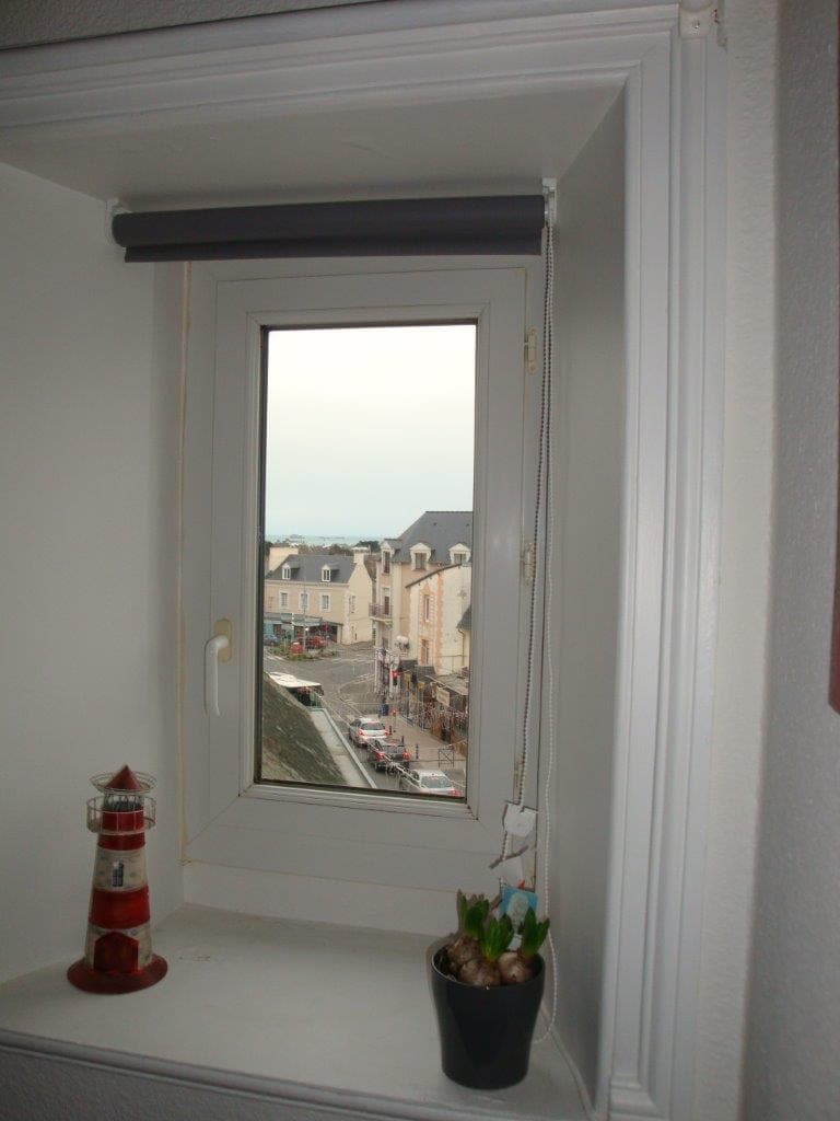 A small window is framed by white trim, offering a view of a quiet street below. A lighthouse figurine sits on the sill beside a potted plant, while the gentle hues of the sky hint at the nearby sea.