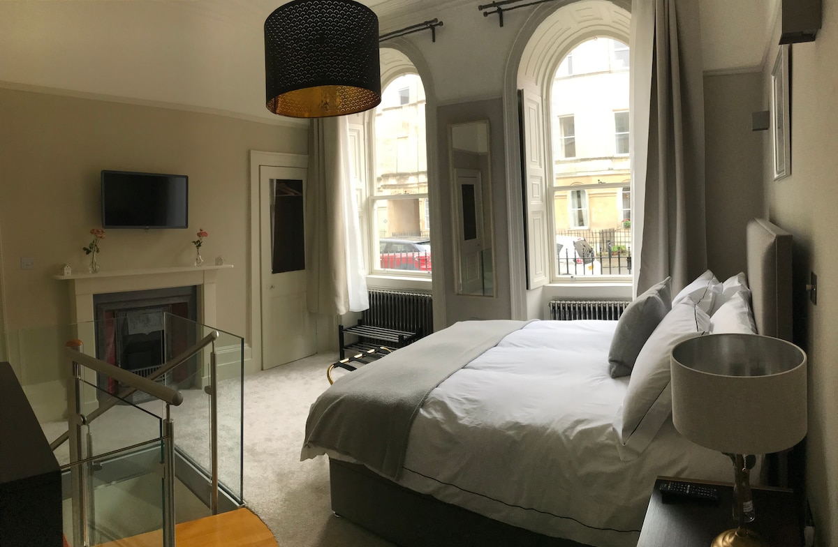 A spacious bedroom features a king size bed with plush bedding, an elegant light fixture, and a large flat-screen TV mounted on the wall. Two tall windows with period shutters provide ample natural light, overlooking the street below. Decorative elements include a tastefully restored fireplace.