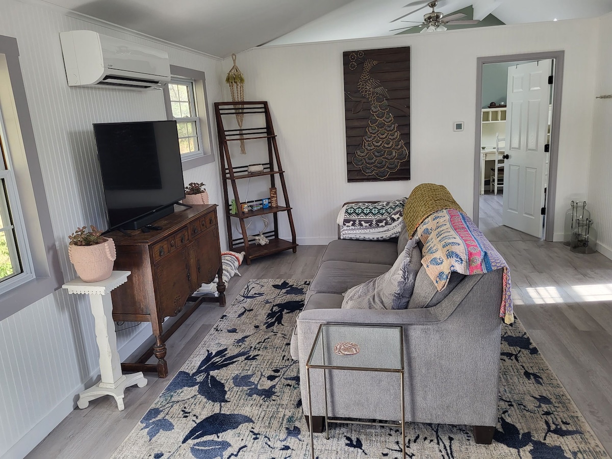 The living area features a comfortable gray sofa adorned with a colorful throw blanket. A wooden console table holds a potted plant, and a wall-mounted flat-screen television sits across from an open bookshelf. Natural light floods the space through the windows.