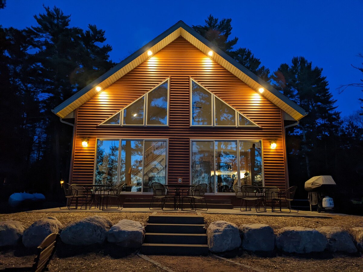 The cabin is prominently featured with a striking A-frame design and large front windows. Evening lights softly illuminate the exterior, accentuating the inviting atmosphere. A stone pathway leads up to the entrance, bordered by a spacious outdoor seating area and a gas grill.