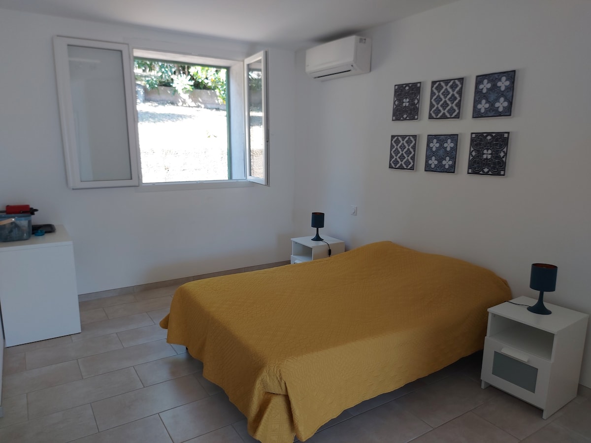 A spacious room features a large bed covered with a yellow blanket. Two nightstands with blue lamps are positioned beside the bed. A window allows natural light to enter, and decorative wall art complements the simple, modern decor.