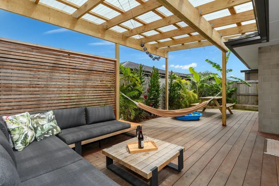 A covered outdoor lounge area is furnished with a spacious sectional sofa adorned with decorative pillows. A wooden coffee table sits centrally, and a hammock is suspended nearby among lush greenery, creating a relaxing space surrounded by nature.