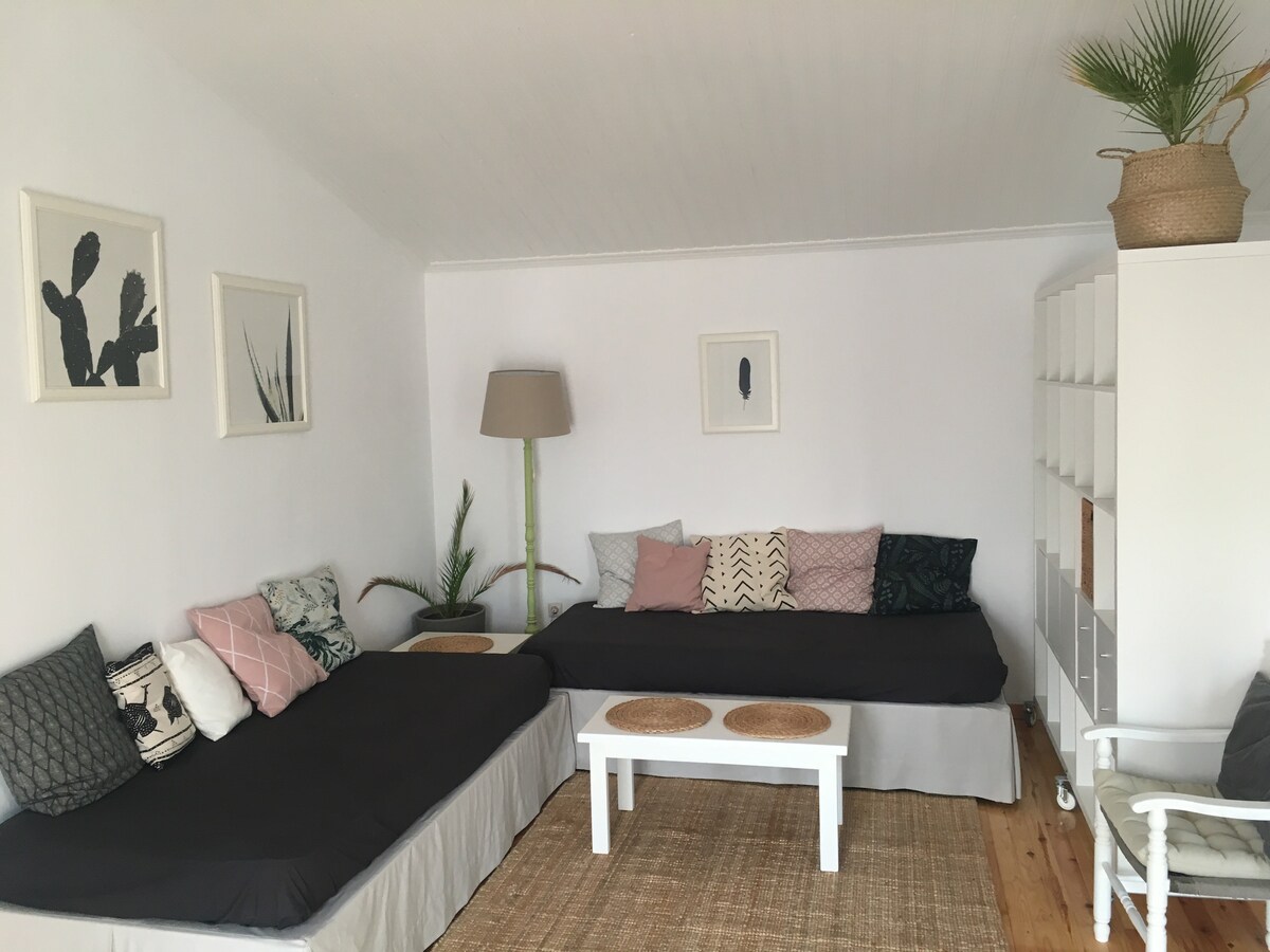 A spacious living area features two large beds with dark covers, complemented by an arrangement of patterned pillows. A light wooden table sits between them, and a woven rug enhances the natural decor. Wall art and a floor lamp add to the room's inviting simplicity.