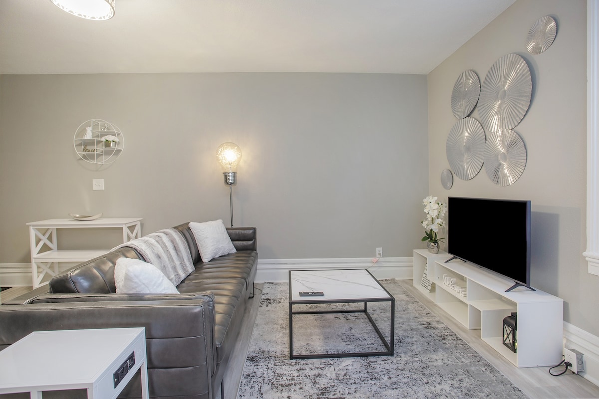 A modern living area features a sleek, dark leather sofa paired with light decorative pillows. A glass coffee table sits in front, while a mounted television is positioned on a white media unit. Artistic metal wall decor complements the neutral-colored walls.