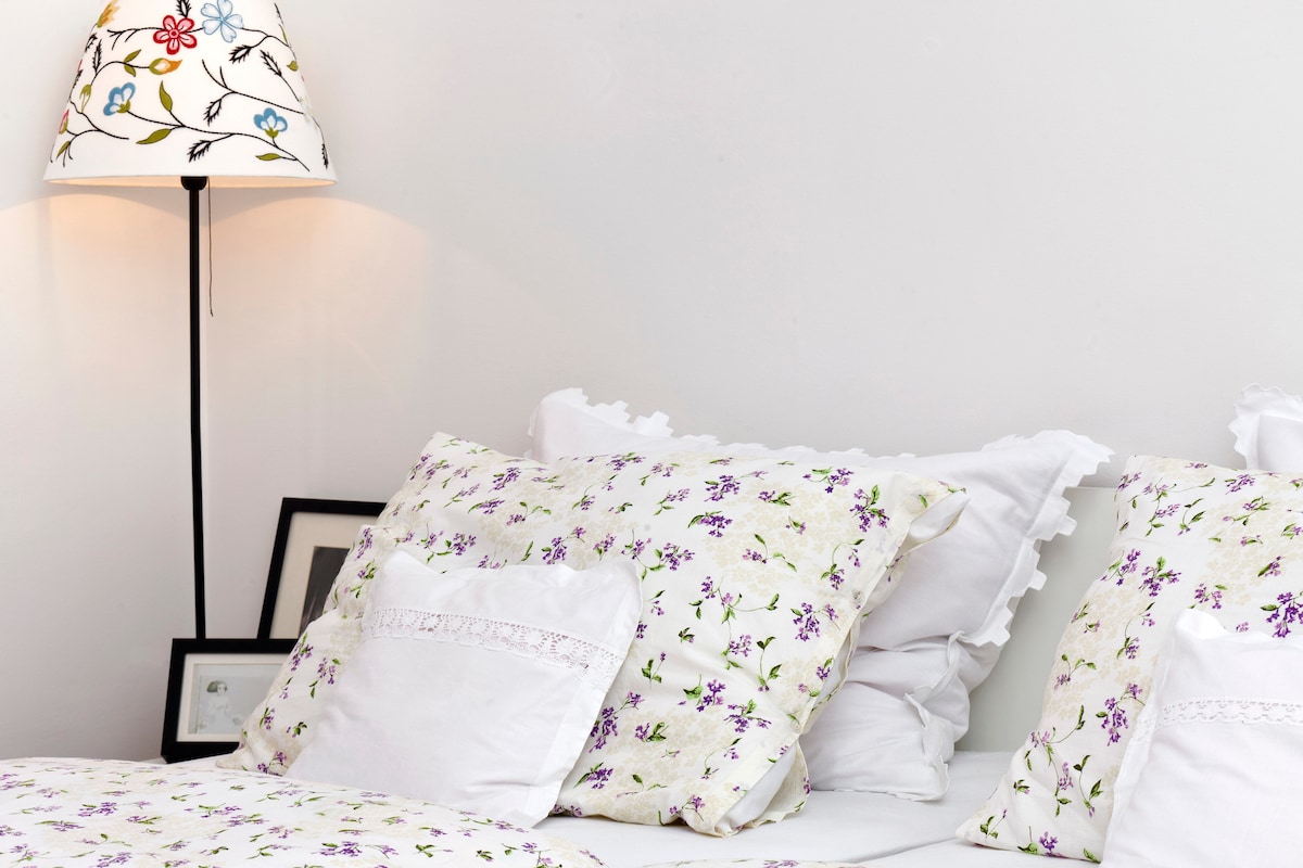 A cozy sleeping area is characterized by a spacious bed adorned with floral-patterned pillows and soft white linens. A decorative lamp with a floral design illuminates the space, while a small collection of framed pictures is subtly positioned nearby.