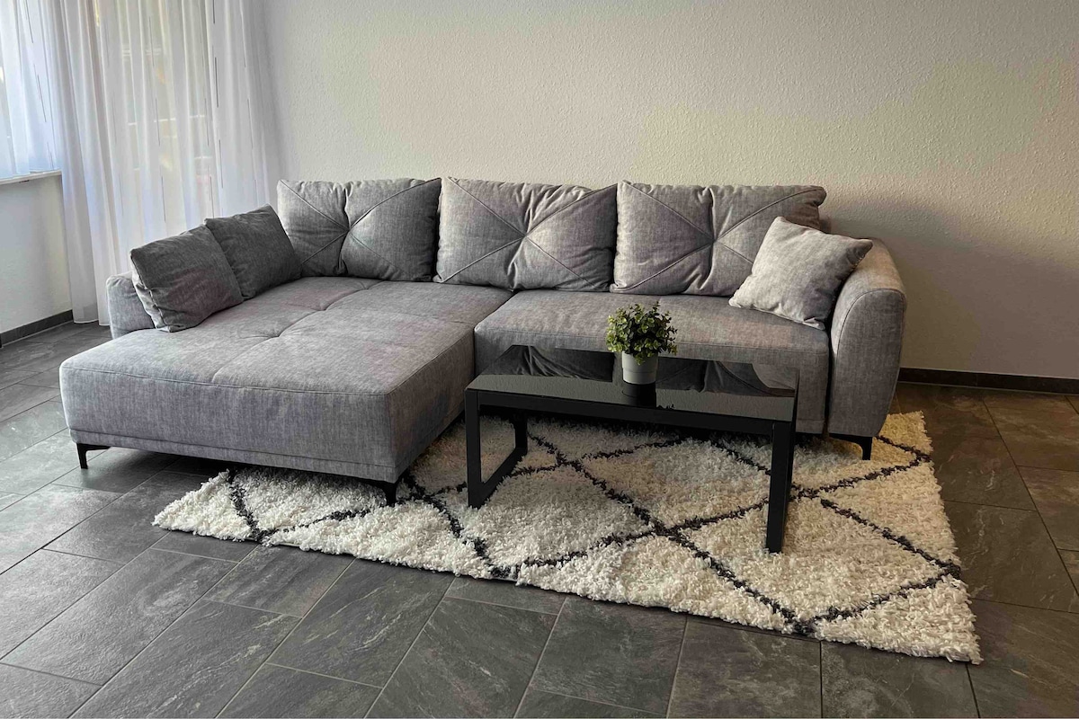 A modern living area features a gray sectional sofa with plush cushions, positioned on a textured rug. A black coffee table sits in front, complementing the open space illuminated by natural light from the nearby window.