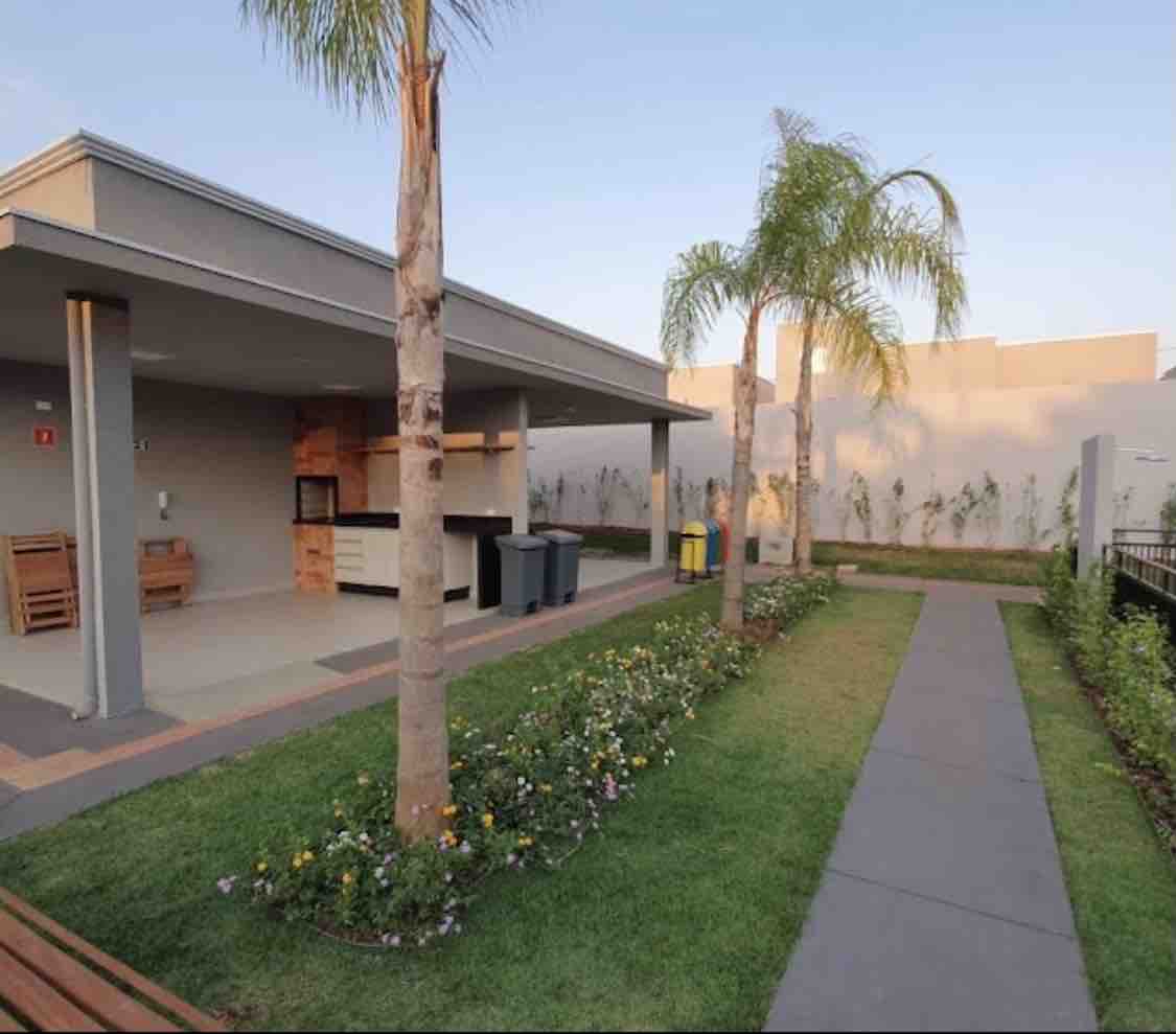 An outdoor gourmet area is showcased, featuring a covered space with a built-in grill, seating, and a small countertop. Lush greenery and blooming flowers line the path, while neatly arranged trash bins are positioned nearby. Palm trees add a tropical touch to the serene environment.