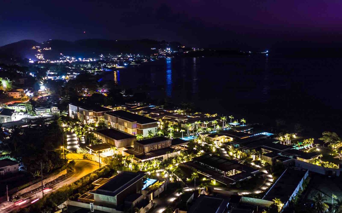 An aerial view captures the vibrant lights of the coastal community at night. The layout of the buildings is highlighted, with pathways illuminated by soft lighting. The water reflects the twinkling lights, contributing to a serene ambiance across the landscape.