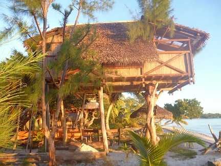 A unique elevated bungalow, supported by wooden stilts, is surrounded by lush greenery. The thatched roof provides a rustic charm, while the spacious terrace below offers dining options in a natural setting by the lagoon. Sunlight illuminates the area, enhancing the coastal atmosphere.