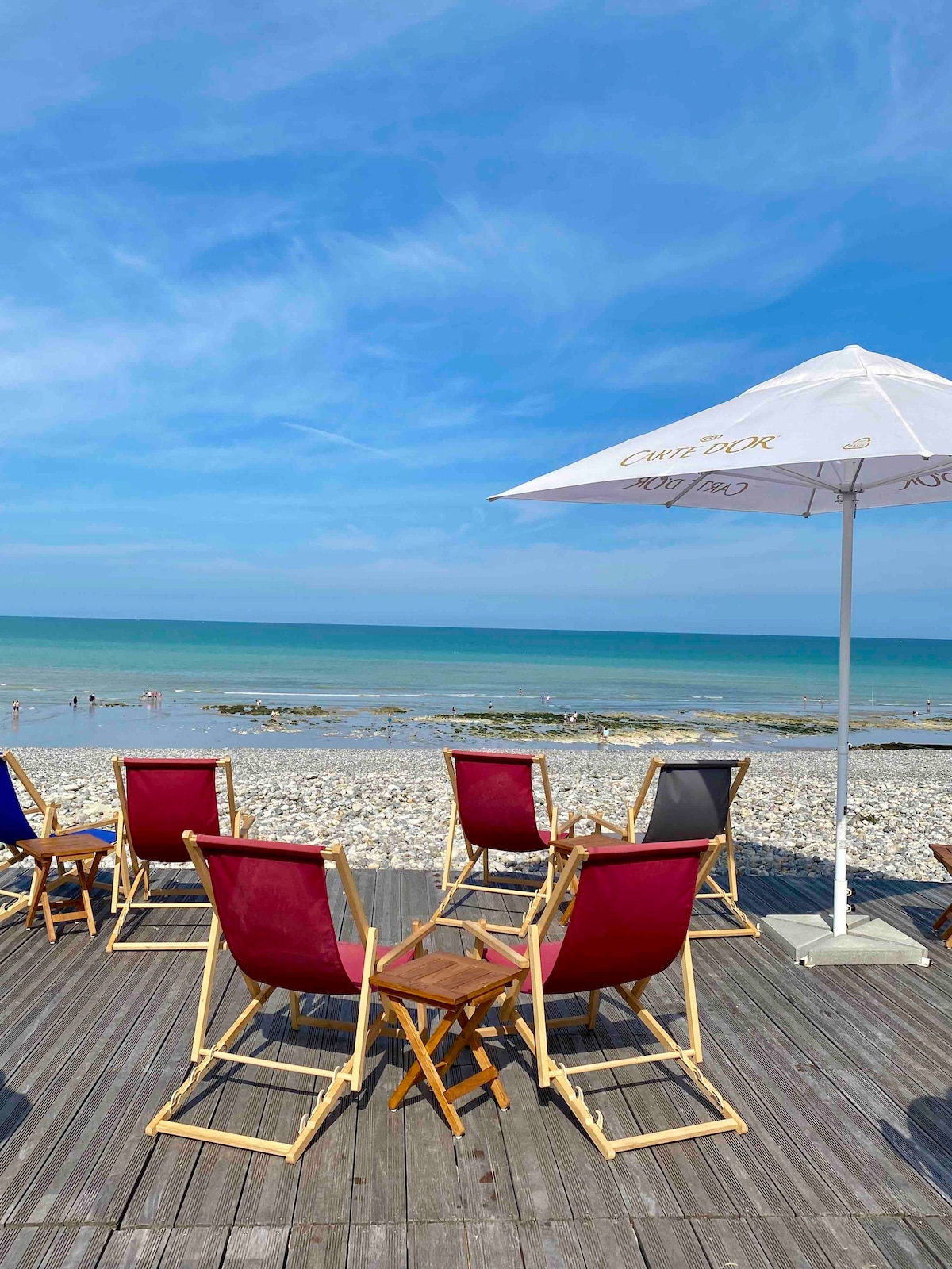 A terrace features several folding chairs in various colors set on wooden decking. An umbrella provides shade, while the beach and calm sea are visible in the background. The sky is clear with soft, scattered clouds, and people can be seen enjoying the water.