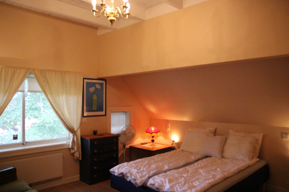The room features a spacious bed with plush bedding, flanked by bedside tables and a cozy lamp. A window with soft drapes allows natural light to illuminate the cream-colored walls. A small dresser and a comfortable seating area add to the inviting atmosphere.