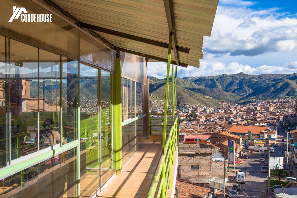 A spacious terrace features wide glass panels, offering expansive views of the surrounding mountains and the city below. Sunlight brightens the area, highlighting the green accents of the railing and walls, creating an inviting space for relaxation.