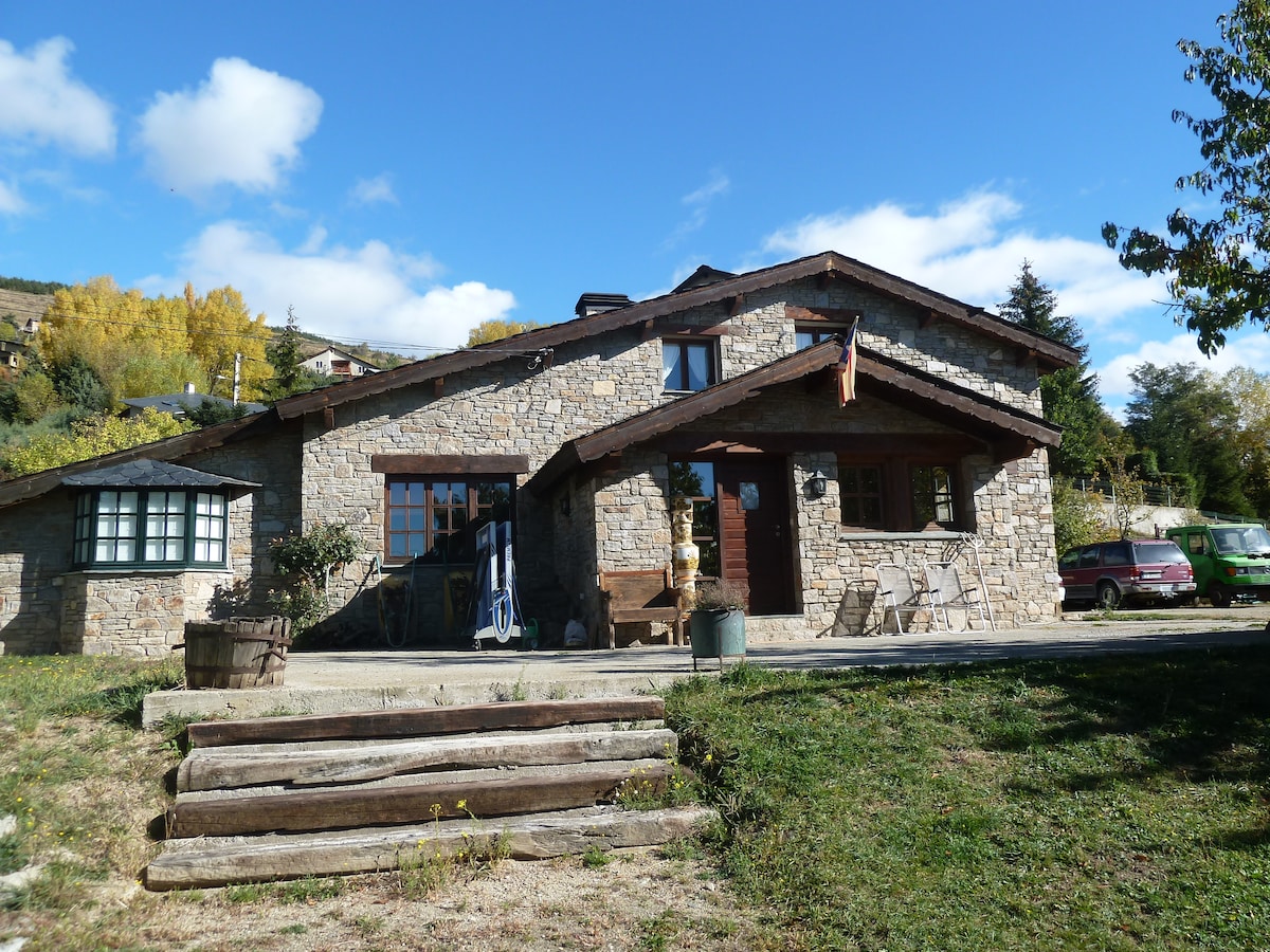 The rustic stone facade of a charming house is framed by a clear blue sky and scattered clouds. Wooden steps lead to a front patio, where outdoor furniture is arranged. Lush greenery and distant trees add to the serene outdoor setting.