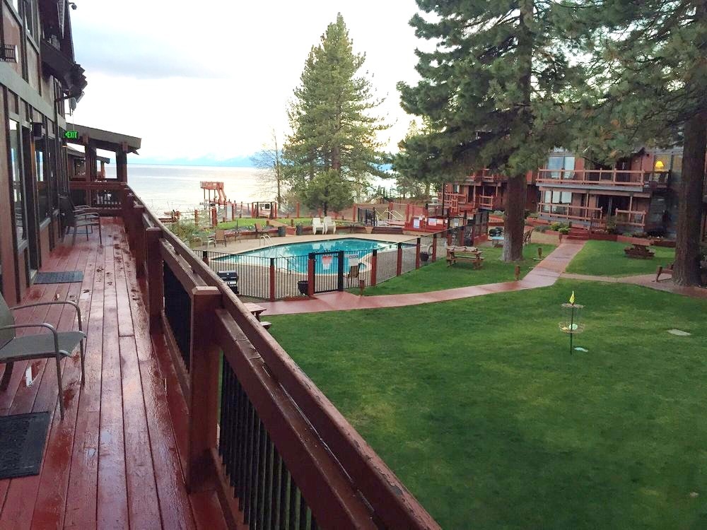 A spacious outdoor area features a seasonal heated pool surrounded by manicured lawns and tall trees. A wooden deck leads toward the pool, with tables and seating arranged on the grass, providing a relaxed atmosphere for enjoyment in the sun.
