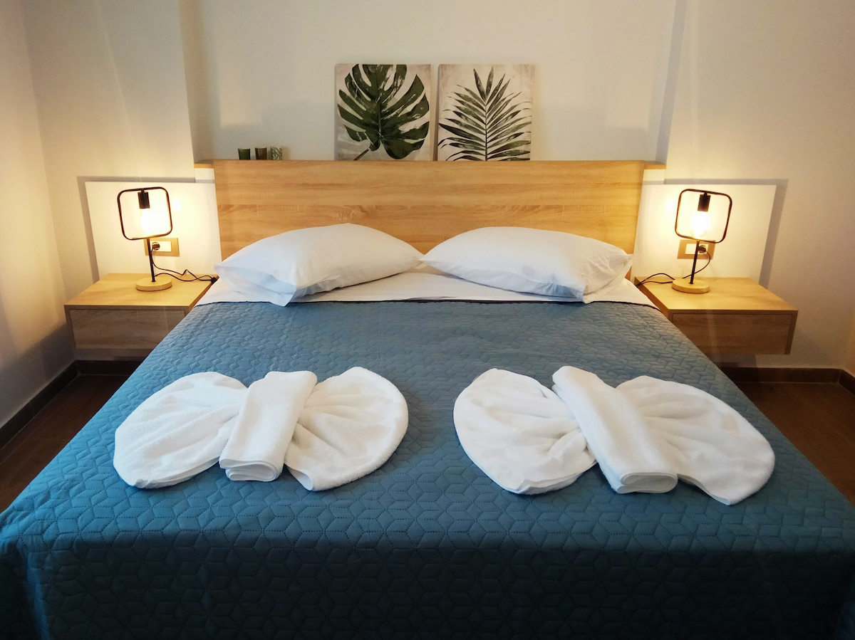 A spacious bed is centered in the room, adorned with a textured blue coverlet and topped with two neatly arranged towels. Two bedside lamps flank the bed, providing soft illumination, while leaf-patterned artwork hangs above the headboard, contributing to a serene ambiance.