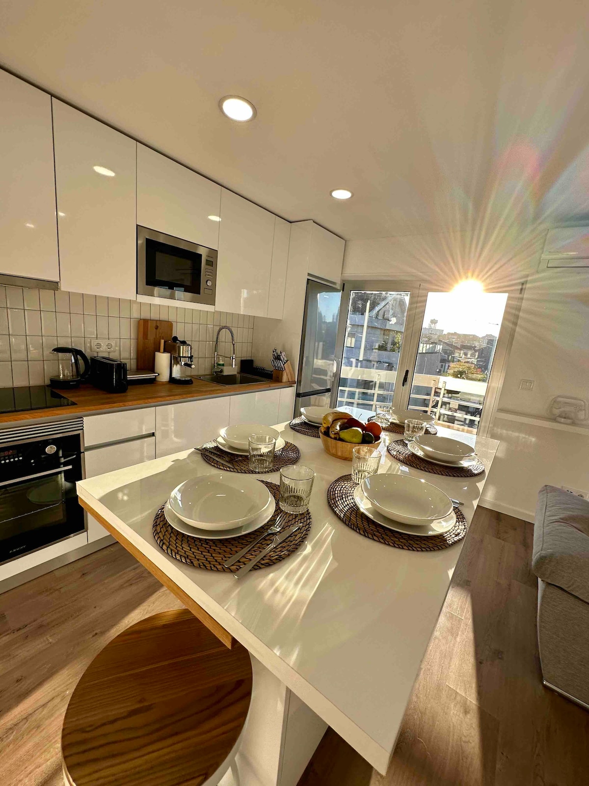 A modern kitchen with sleek white cabinets features a breakfast bar set for four, complete with plates and glasses. A microwave and oven are integrated into the cabinetry. Light streams in through the balcony doors, highlighting the fresh fruit centerpiece on the table.