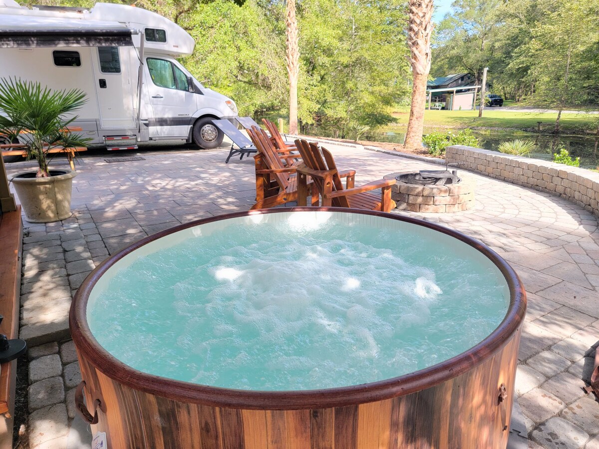A wooden hot tub filled with bubbling water is centered in the outdoor space. Adirondack chairs are arranged around a fire pit nearby. In the background, a recreational vehicle is parked under trees, complementing the serene setting.