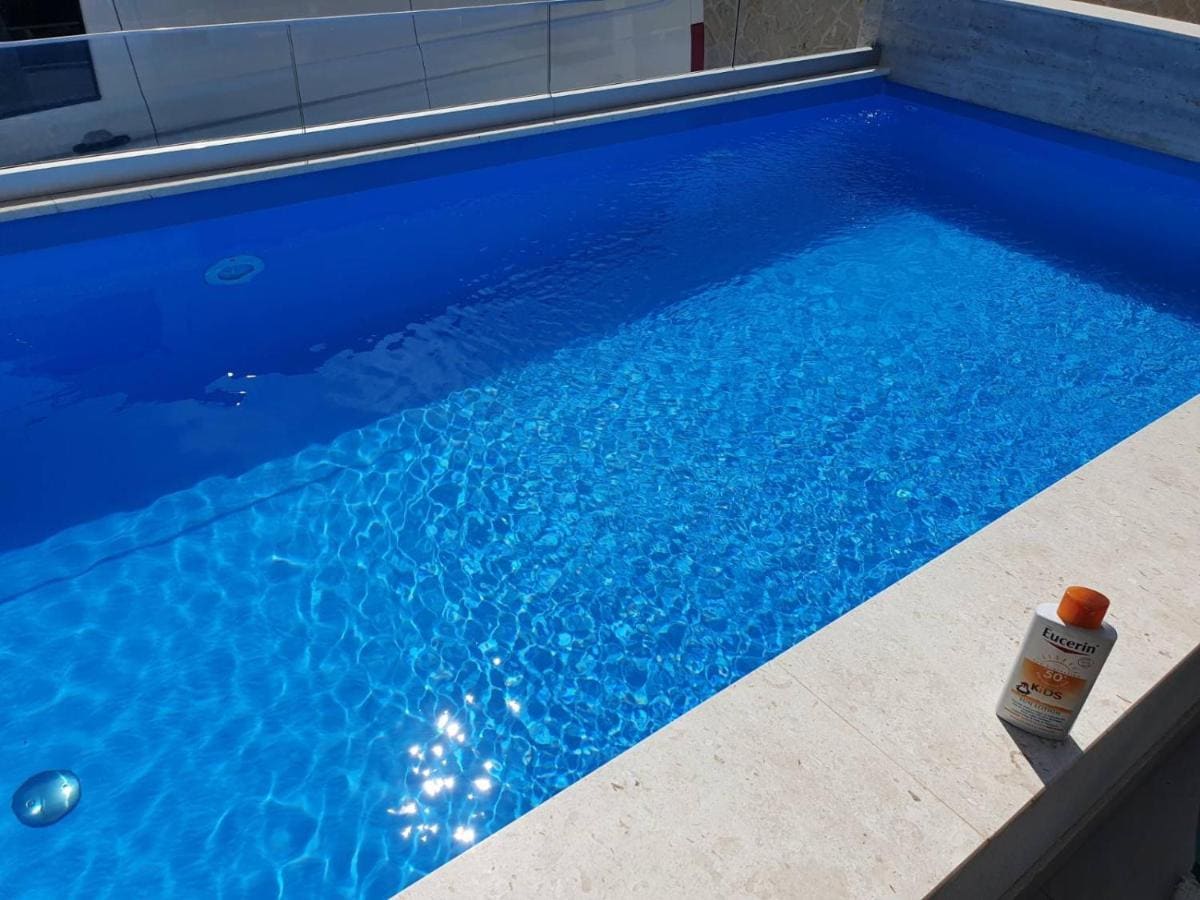 A private outdoor heated swimming pool features clear blue water reflecting sunlight. The pool's edge is lined with light-colored stone. A bottle of sunscreen is placed on the edge, adding a touch of relaxation to the serene aquatic setting.