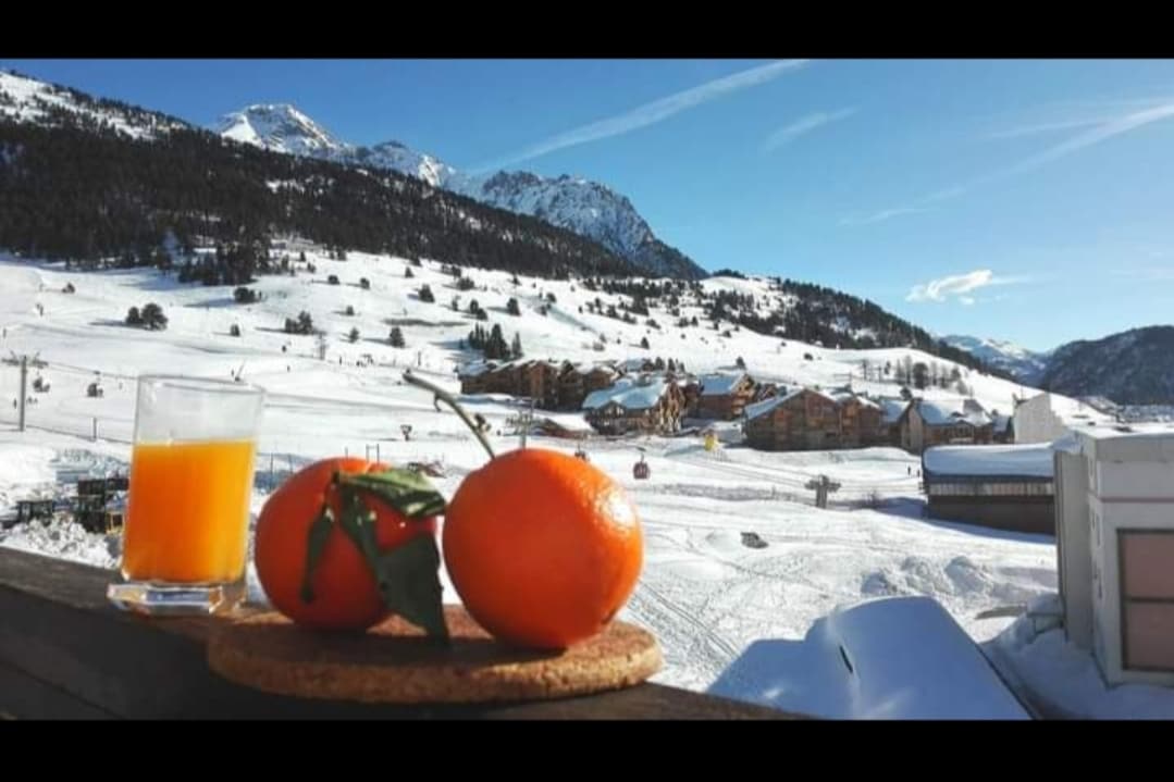 A refreshing view from a balcony showcases a snowy landscape with mountains in the background. A glass of orange juice and two oranges are displayed on a wooden coaster, adding a touch of color to the scene. The ski slopes and buildings are visible below.