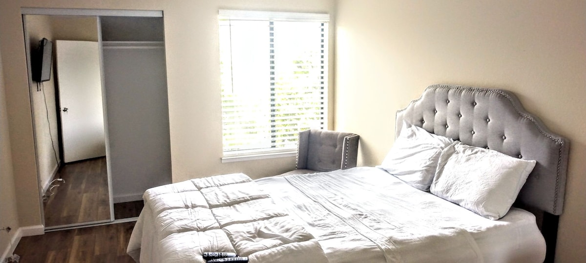 A serene sleeping area features a spacious bed with crisp white bedding positioned near a window lined with blinds. A plush chair sits in the corner, and a mirrored closet door reflects the soft natural light in the room.