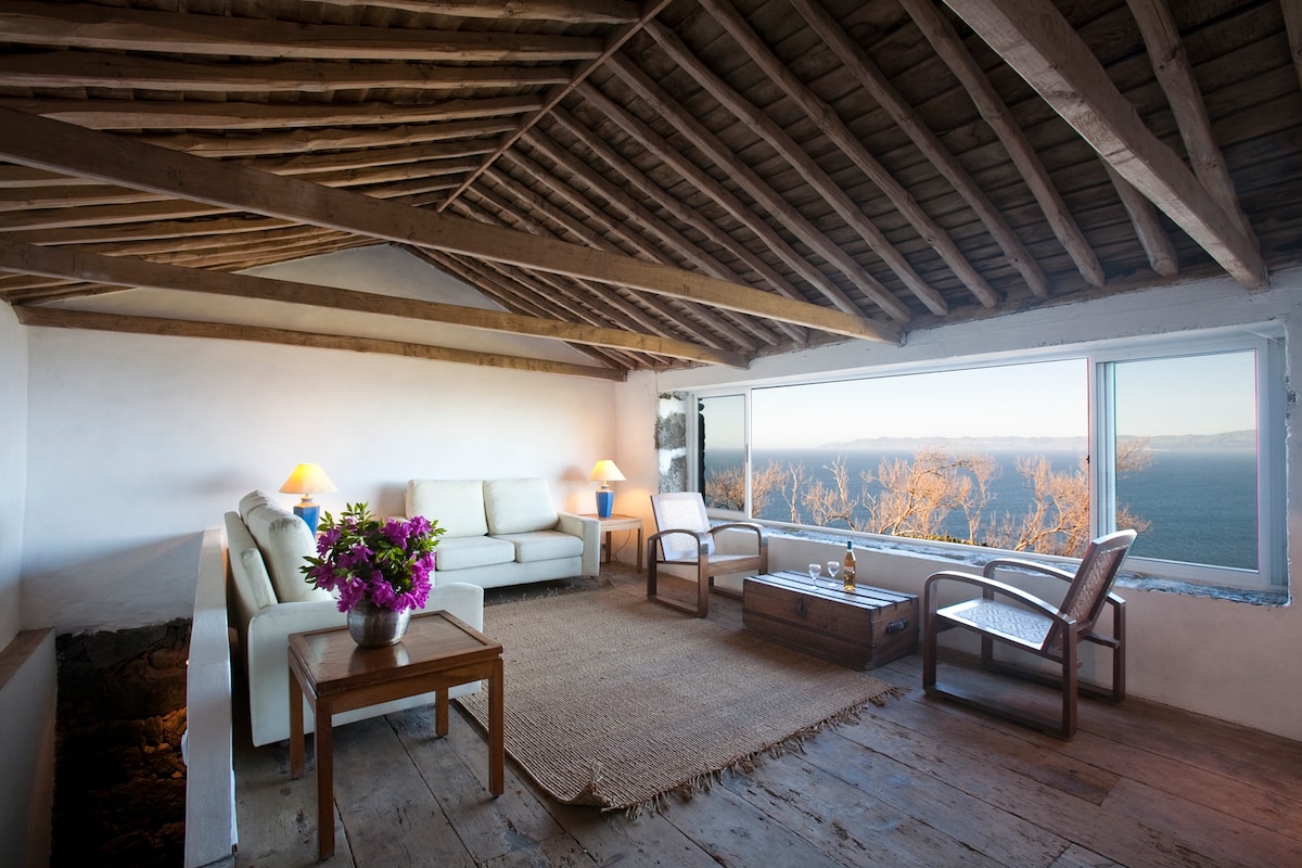 A spacious lounge area features comfortable seating with soft cushions, complemented by a wooden coffee table. Large windows provide panoramic views of the landscape, while a woven area rug adds texture to the wooden floor. Fresh flowers adorn the table, contributing to the room's welcoming ambiance.
