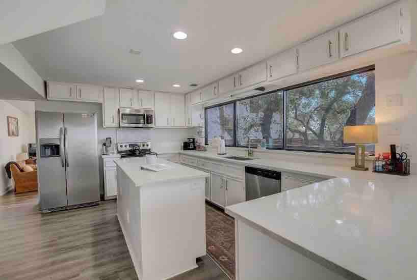 A modern kitchen is presented with white cabinets and stainless steel appliances. Natural light enters through large windows, offering a view of surrounding trees. An island with a countertop serves as a functional workspace, while a lamp adds a touch of warmth to the area.