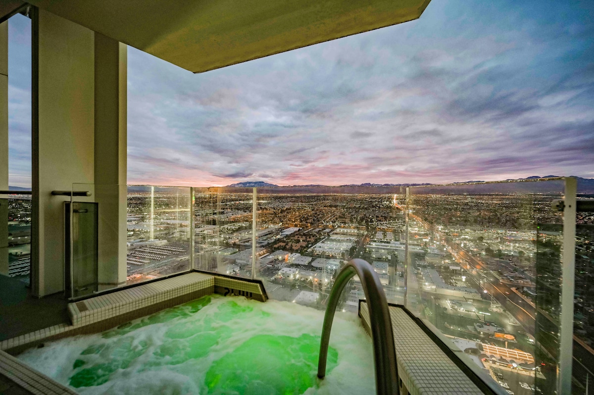 A private Jacuzzi tub sits on the balcony, offering sweeping views of the city below and distant mountain ranges. The tub is surrounded by glass railings, enhancing the serene atmosphere during sunset.